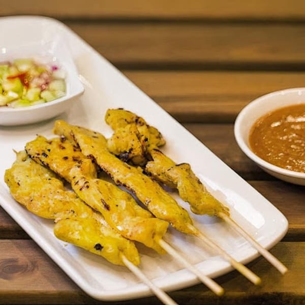 Chicken satay skewers with peanut sauce and cucumber relish on a white plate.